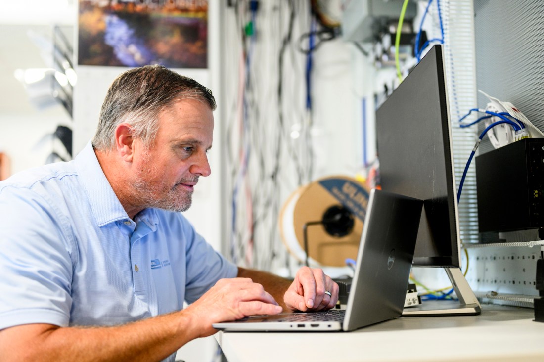 Joe Ranahan wearing a collared shirt leaning over a computer working on a program to display lights on the Boston campus buildings.