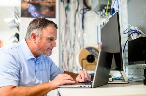 Joe Ranahan wearing a collared shirt leaning over a computer working on a program to display lights on the Boston campus buildings.