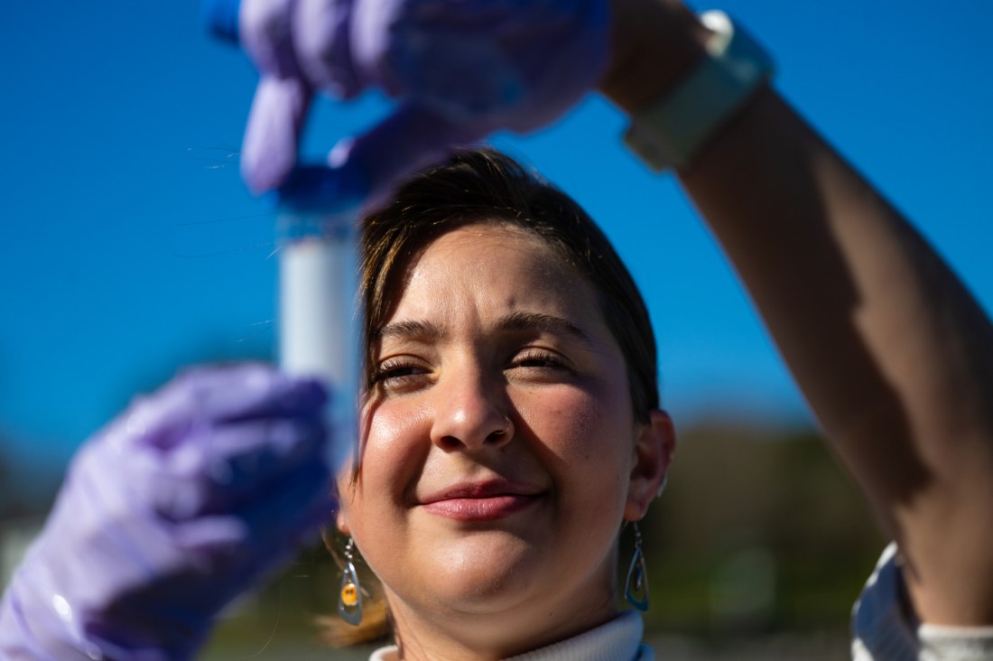 A woman wearing purple gloves examines water samples against a blue sky.