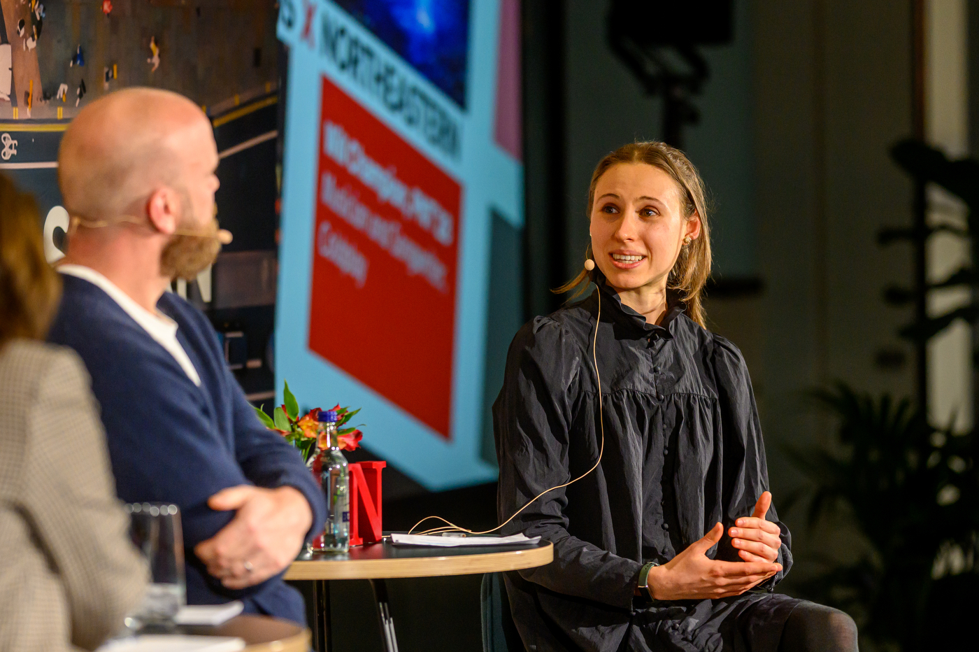 A person wearing a black shirt and a microphone speaking to Will Champion on stage during a panel discussion. 
