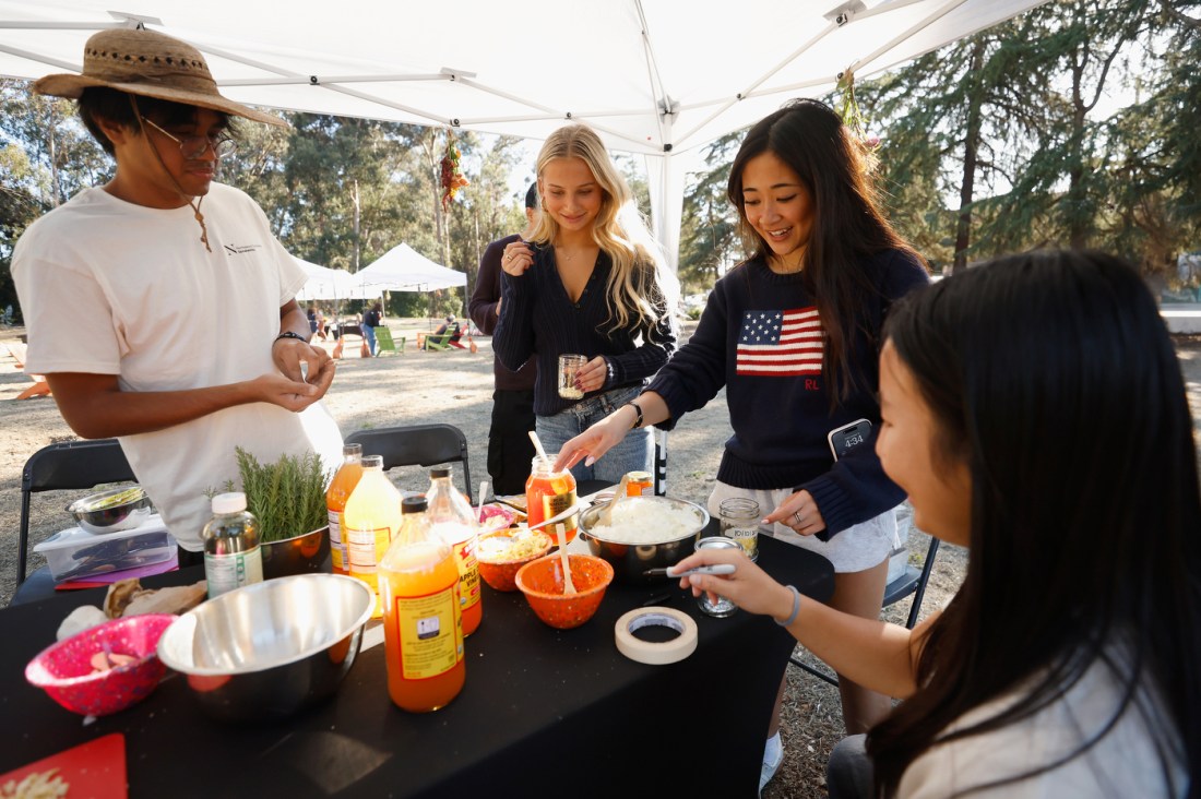 Four people stand around a table with a black tablecloth that has various foods on it, including a large jar of honey.