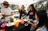 Four people stand around a table with a black tablecloth that has various foods on it, including a large jar of honey.