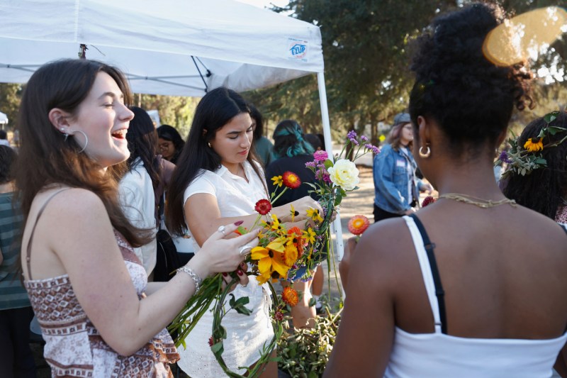 People browse colorful flower arrangements at an outdoor market under a white tent canopy. In the foreground, a person with their hair in a bun views the display from behind, while two young women in white tops examine bouquets featuring yellow, orange, red, and white blooms. Trees and additional visitors are visible in the sunny background.