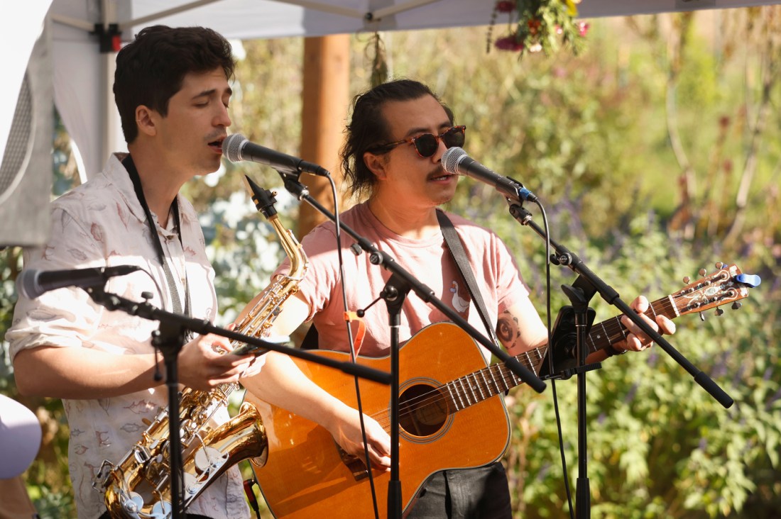 Two musicians singing into microphones, one holding a saxophone and one holding a guitar.