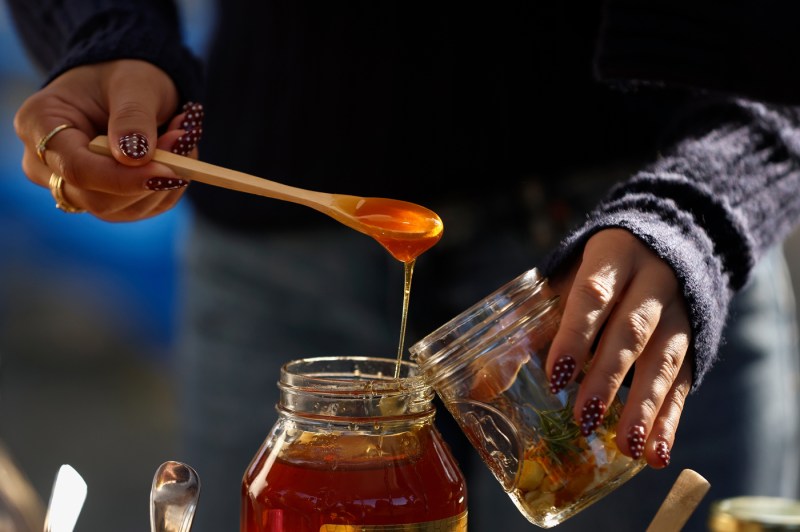 A close-up shows hands drizzling golden honey from a wooden dipper into a glass jar. The person wears multiple gold rings and has decorated nails with a dotted pattern.