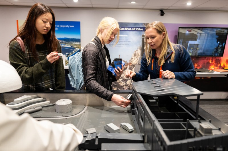 Three people gather around a tabletop demonstration in an exhibition space, closely examining what appears to be a miniature architectural model or simulation. They are all focused intently.