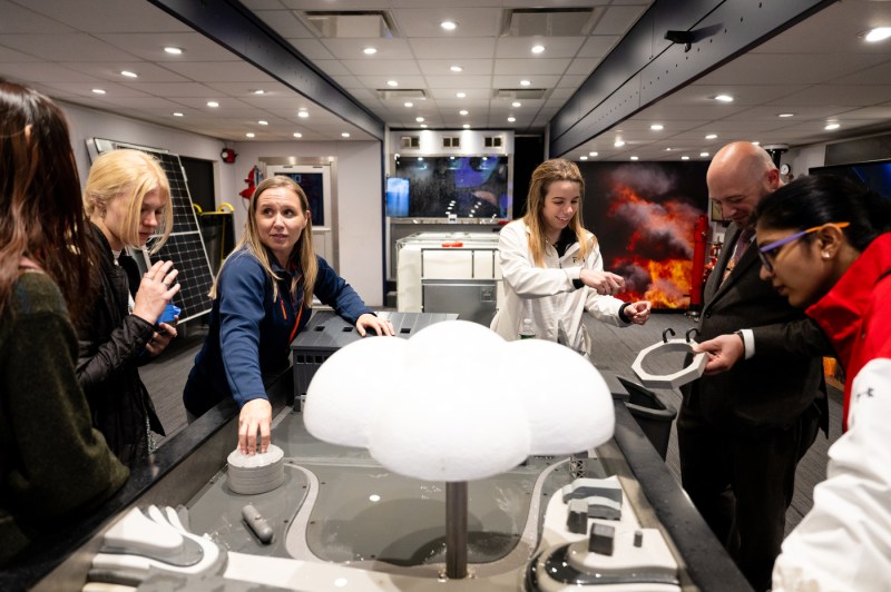 A group of people examines an interactive display inside of a modern exhibit space. The display features a white cloud-shaped unit over a tub or basin. 