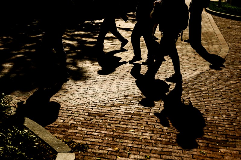 A ground-level view captures silhouettes of students' legs and the shadows they cast on brick pavement as they walk at what appears to be sunset. 