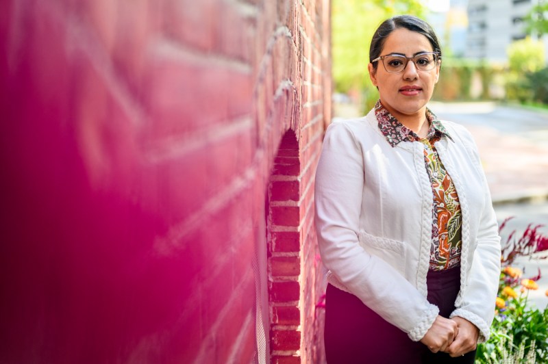 A woman with dark hair pulled back and a white coat over a patterned blazer poses against a brick wall on a sunny day. 