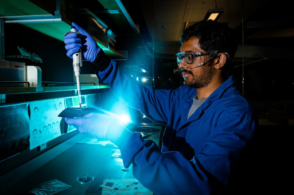 A biotechnology student wearing blue lab coat, protective gloves, and safety glasses pipettes liquid onto an embroidered surface in a dimly lit laboratory illuminated by blue-green light. The researcher carefully dispenses precise amounts of liquid using a pipette held in one hand while working over what appears to be a textile sample on a lab bench, demonstrating the intersection of traditional craft techniques and modern biotechnology research.