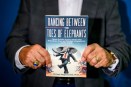 A man holds up the cover to a book titled: "Dancing Between the Toes of Elephants: Clever Private Equity Idioms and Anecdotes from an Outsider’s Experience on the Inside."