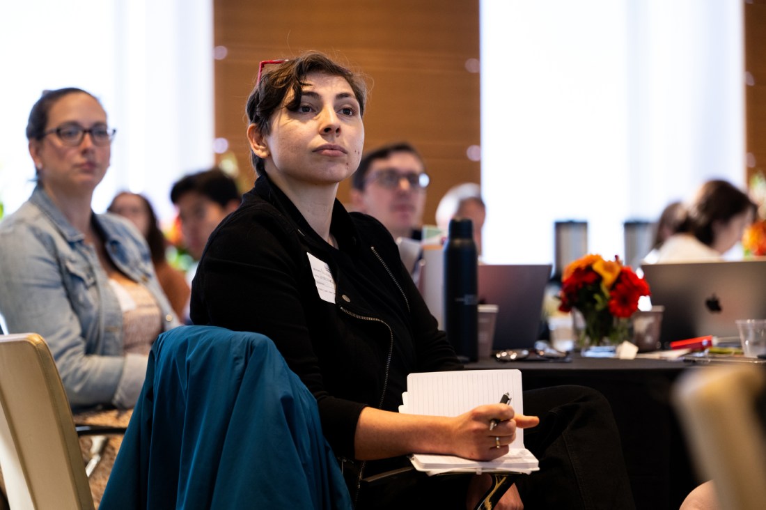 A person wearing a black blazer and red glasses propped on their head sits at a table and listens intently to a panel discussion at the Social Media Summit. They are taking notes in a notebook while listening. 