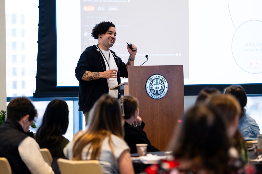 A speaker with curly hair and tattoos on their arms stands at a wooden podium with the Northeastern University seal, gesturing while holding a slide clicker and addressing an audience. Attendees are visible seated in the foreground. 