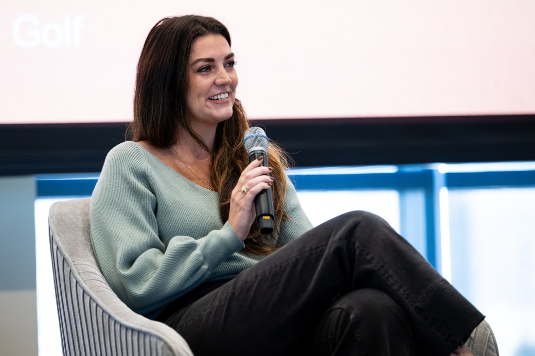 A person with long dark hair smiles while holding a microphone. She is seated on stage and a presentation screen is visible behind her. 
