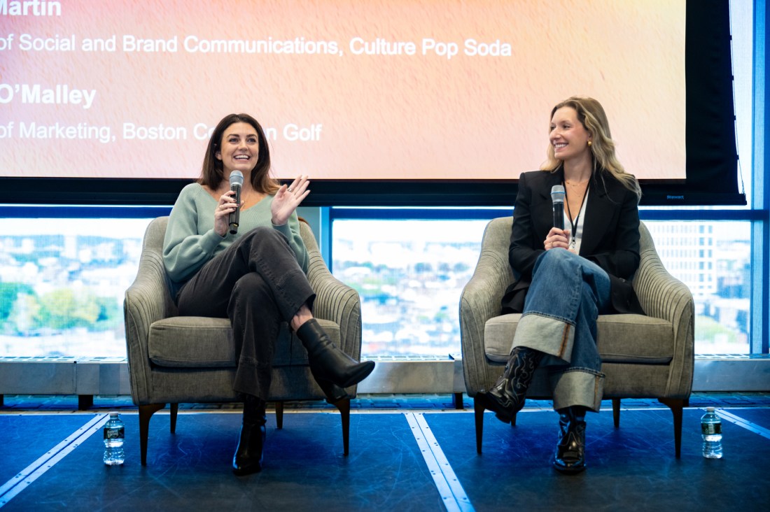 Eileen O'Malley and Haley Martin sit on stage in grey armchairs in front of a presentation screen and large windows with a city view. They are both holding microphones as they present on a panel at the Social Media Summit. x