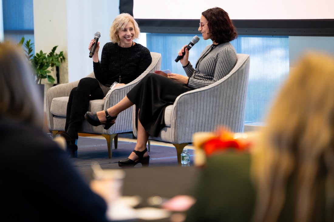 Two women sitting in curved grey chairs on a stage, engaged in a conversation during a panel discussion. They are both holding microphones. Attendees are visible in the foreground. 