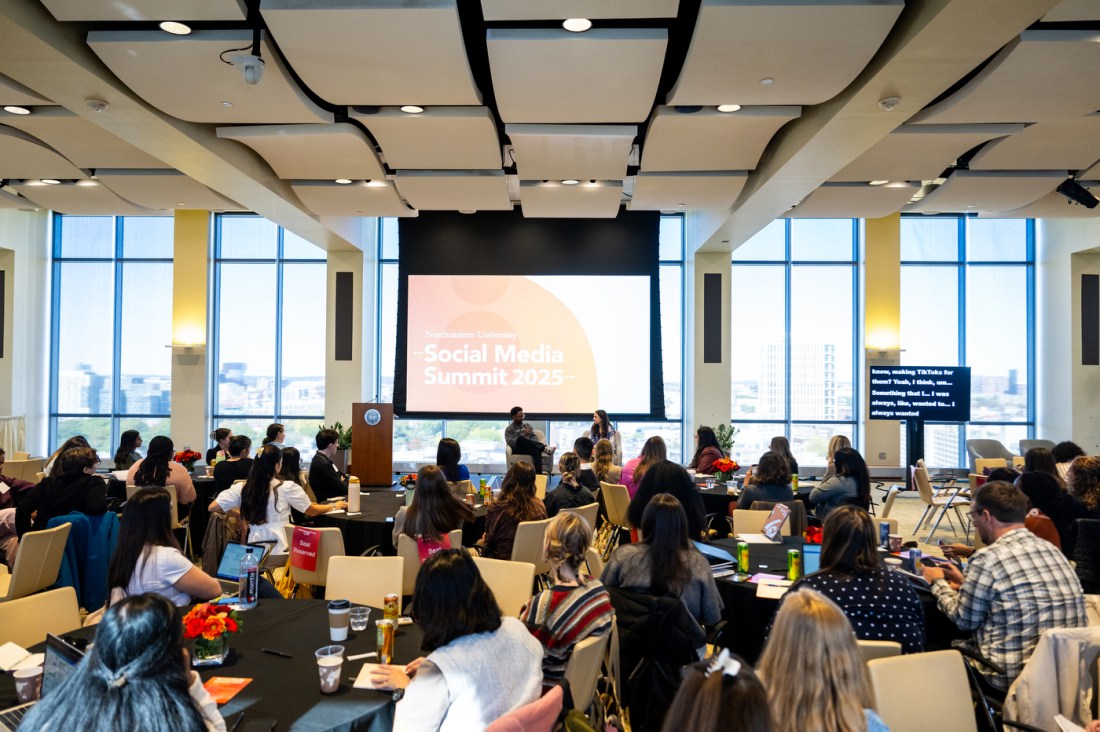 A large conference room with attendees seated at round tables. Large windows line the back wall. 