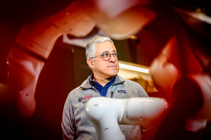 Professor Taskin Padir stands in his robotics laboratory, photographed through red robotic components in the foreground that frame the image, wearing glasses and a gray fleece vest.