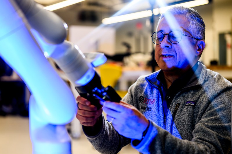 Taskin Padir, wearing glasses, examines a white robotic arm in a laboratory setting with blue-purple lighting. 