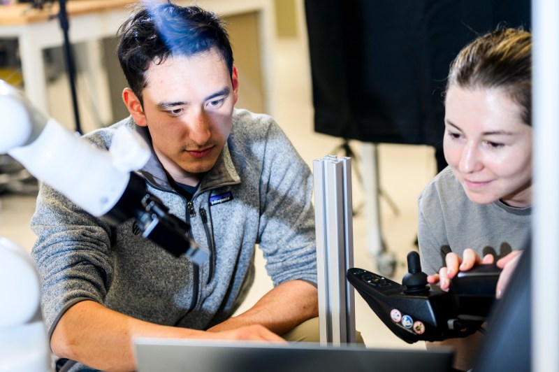 Drake Moore and Damla Leblebicioglu work together, focused on assembling or adjusting a robotic arm attachment for a wheelchair. 
