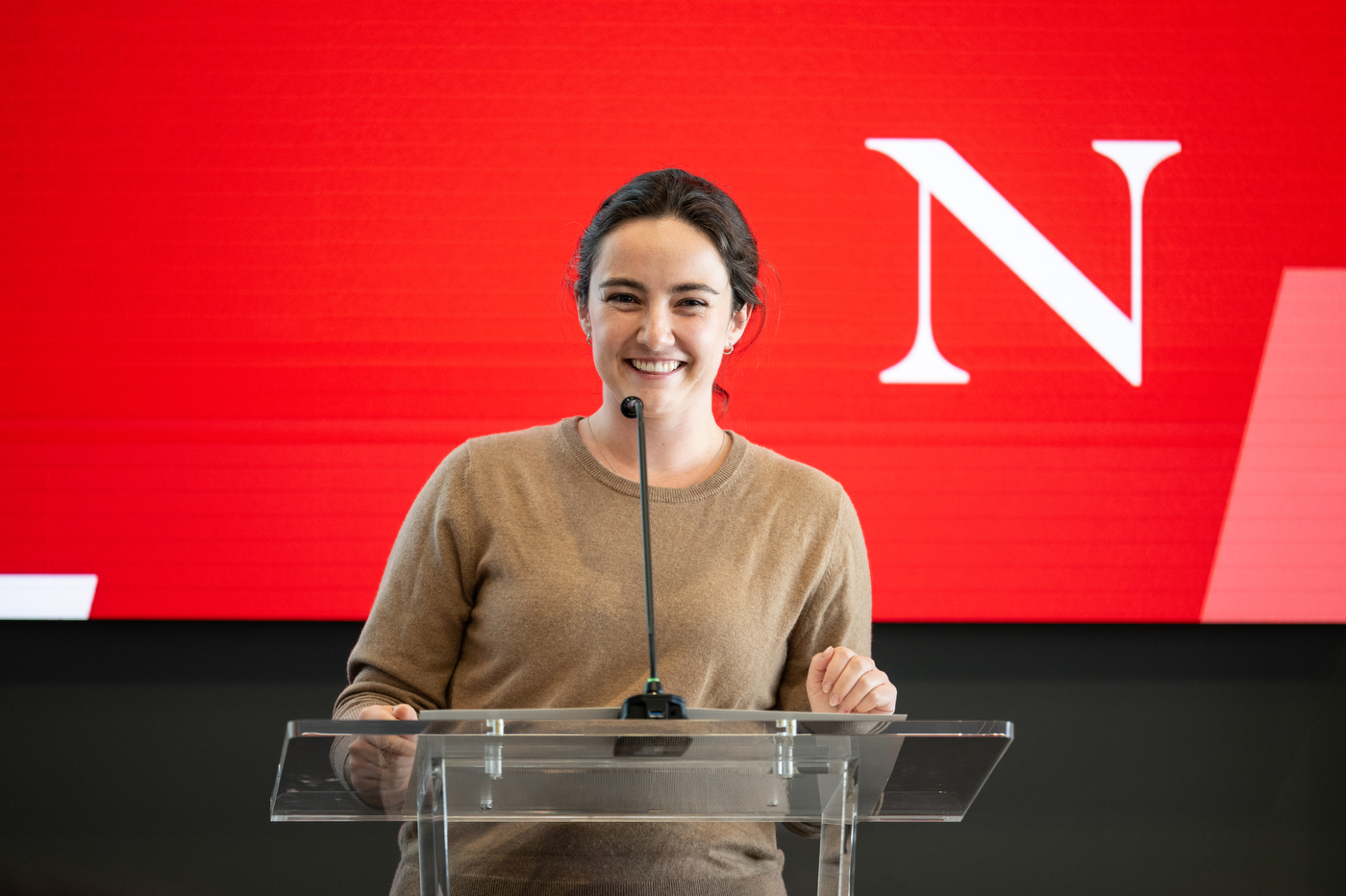 Northeastern University student smiles at a podium with a red backdrop featuring the Northeastern “N” logo.