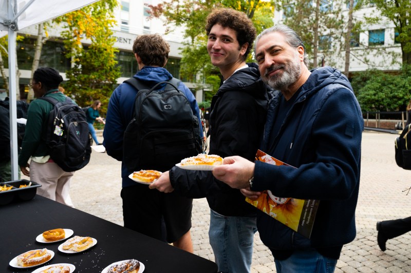Two men smile while holding plates of pastries at an outdoor campus food event, with more pastries displayed on a table in the foreground and students gathering in the background.