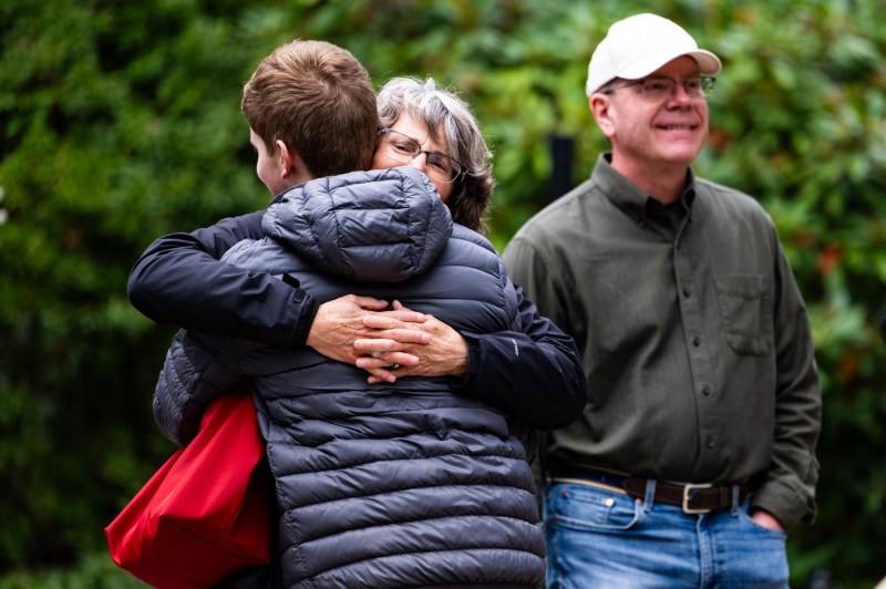 A woman with gray hair and glasses embraces a person in a navy blue puffy jacket, shown from behind. The woman's hands are visible clasped around the person's back. To the right, a smiling man wearing a white baseball cap and olive green button-down shirt with jeans stands nearby. A bright red bag is visible on the embracing woman's shoulder. The scene takes place outdoors against a backdrop of lush green foliage.