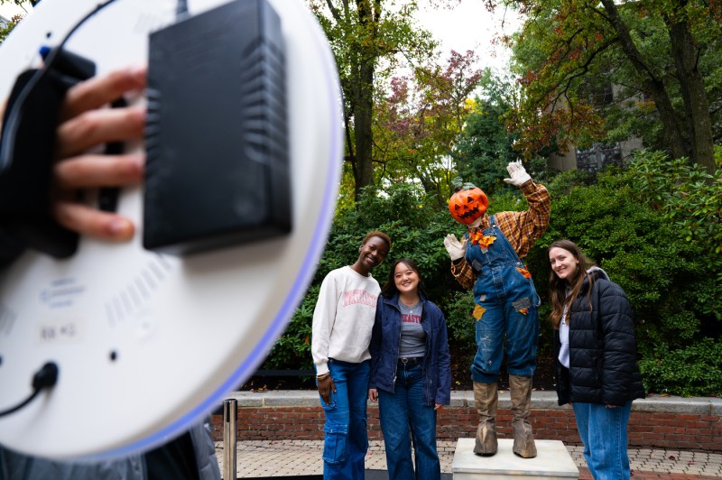 Students pose with seasonal scarecrows including one with a jack-o-lantern head as someone photographs them with a smartphone in the foreground on an autumn campus day.