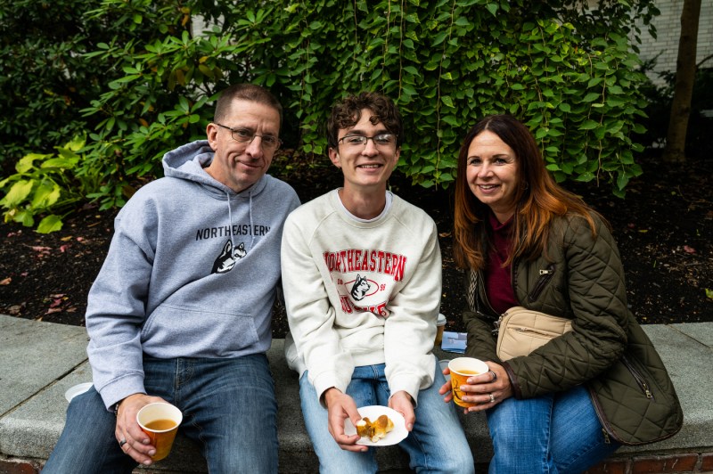 Three people wearing Northeastern University sweatshirts sit together on a bench in front of a lush green ivy wall, smiling while holding drinks and snacks.
