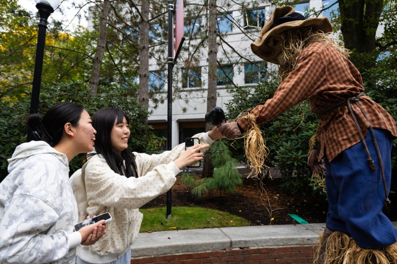 Two students in light-colored jackets reach out to interact with a scarecrow wearing a tan hat, orange checkered shirt, and blue overalls on an outdoor campus lawn.