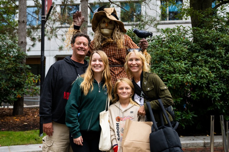 Family of four poses with a straw-hatted scarecrow adorned with a black crow on an autumn campus, holding tote bags and smiling in front of a modern building.
