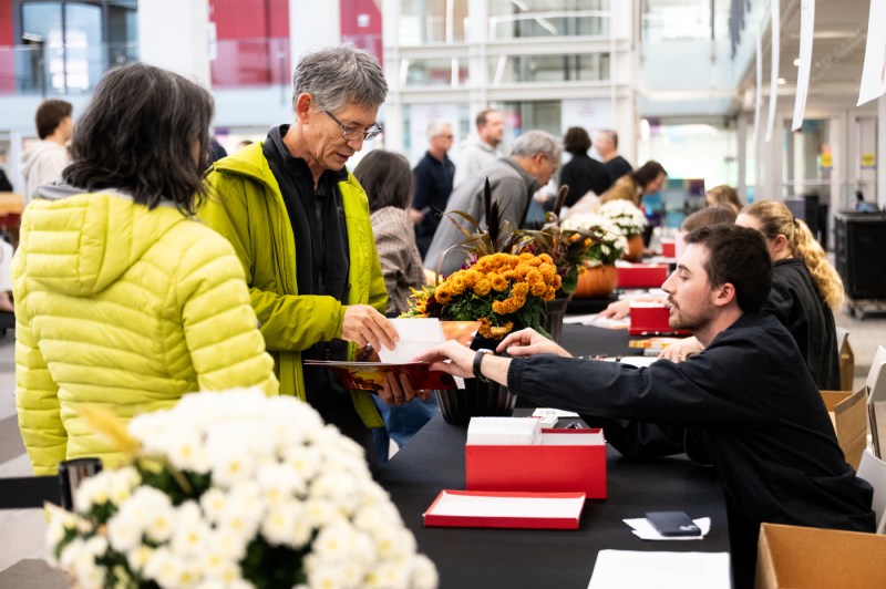 Two visitors in bright yellow jackets receive materials from a student volunteer at a flower-decorated table during a campus event in a modern atrium.