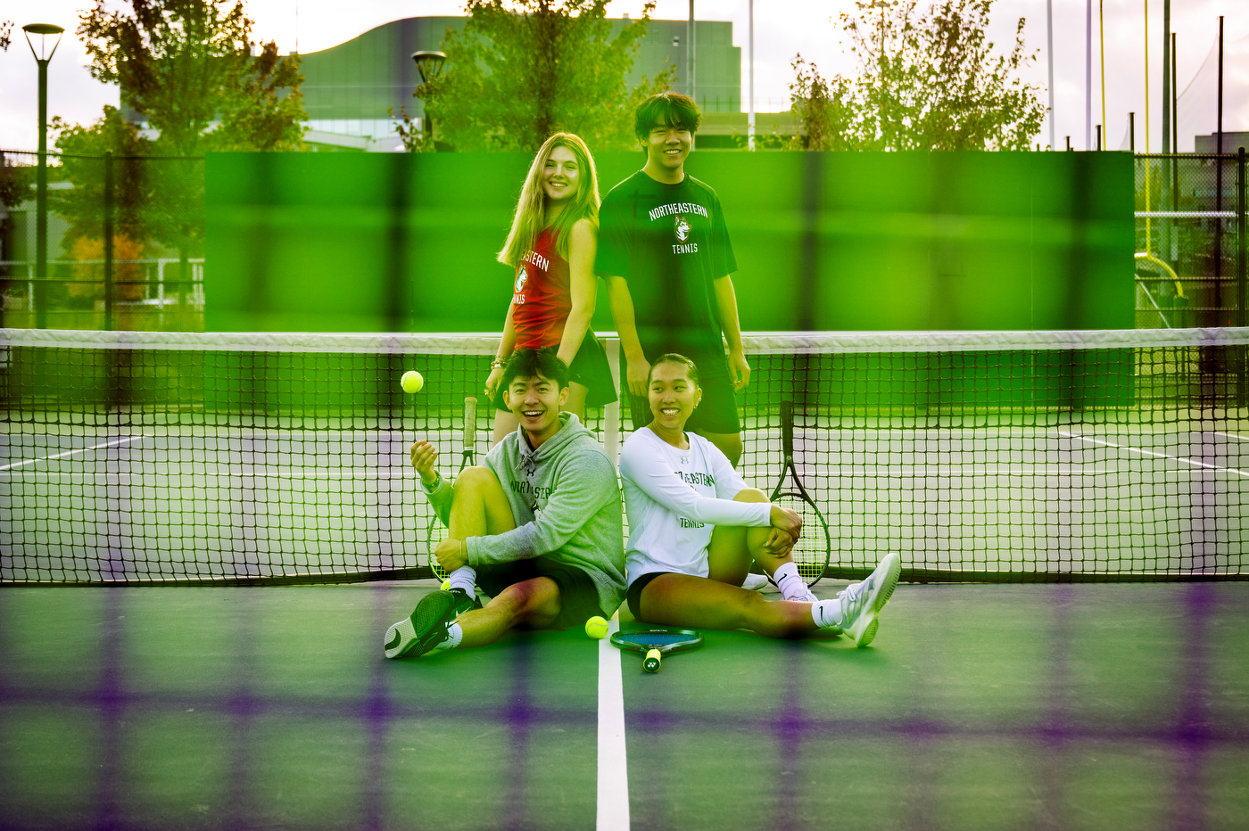 Four members of the Northeastern University tennis team pose together on an outdoor tennis court. In the front row, two players sit cross-legged on the court surface - one holding a tennis ball and wearing a gray Northeastern sweatshirt, the other in a white long-sleeve Northeastern Tennis shirt holding a tennis racket. Standing behind them are two more team members wearing Northeastern athletic wear - one in a red tank top and another in a black Northeastern Tennis t-shirt. The photo is taken through a tennis racket with the people in front of the net with green tennis court 