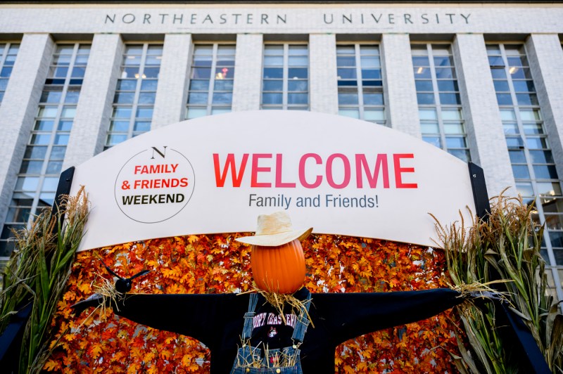 A large “Welcome Family and Friends!” banner with fall decorations stands in front of a Northeastern University building during Friends and Family Weekend.