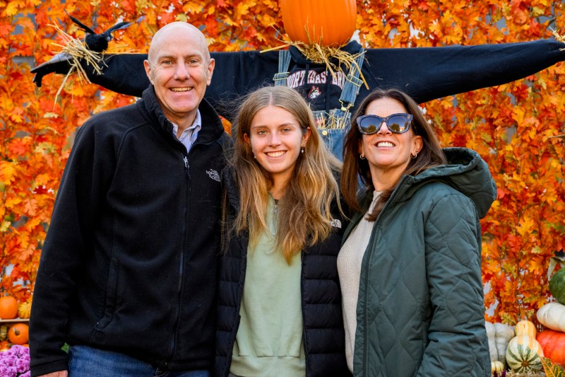 A family of three smiles for a photo in front of autumn decorations and a scarecrow at Northeastern University’s Krentzman Quad during Friends and Family Weekend.