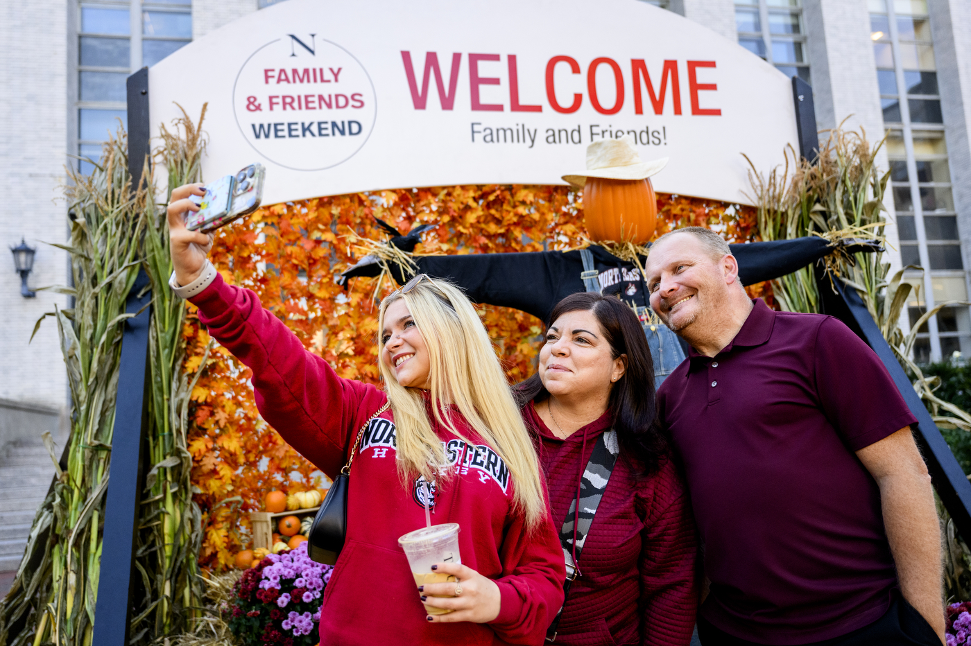A family poses for a selfie in front of a "Welcome family and friends" sign, which also has fall foliage around it. 