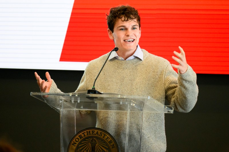 A young person stands behind a podium with a microphone, gesturing while speaking against a red-and-white backdrop.