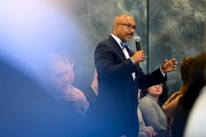 Speaker in a suit holds a microphone and gestures while seated in front of a softly lit backdrop during a fireside-chat event.