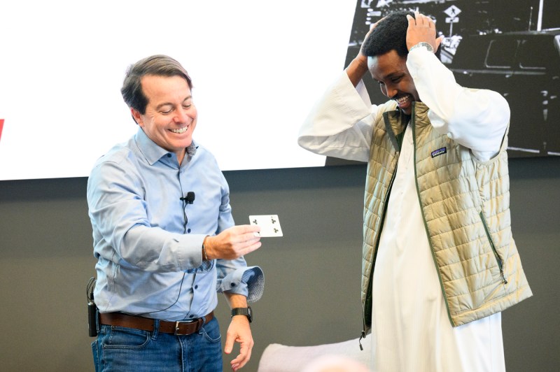Two people laugh and shake hands onstage during a fireside chat event, one adjusting their jacket collar while smiling.