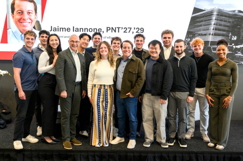 A group of students and President Joseph E. Aoun stand together onstage in front of a large portrait and event signage, smiling for a group photo.