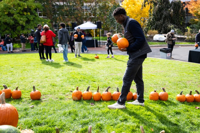 Students and families pick pumpkins on a green lawn surrounded by autumn trees during Northeastern University’s Friends and Family Weekend Fall Festival in Boston.