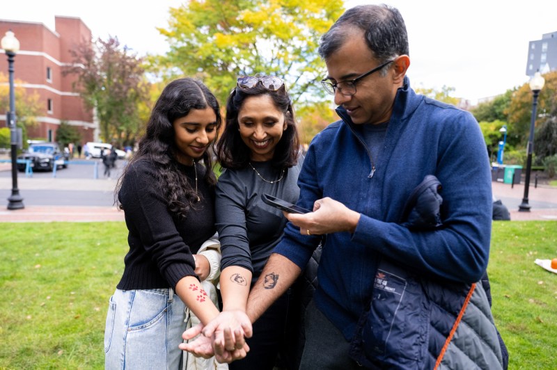 A family of three, a daughter in a black top, a woman in a black jacket center and a man in a blue vest right display tattoos on their inner forearms. They stand with their arms extended showing the tattoo designs as the man takes photograph.