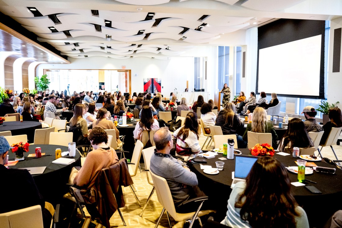 A large conference style room with attendees sitting around round tables. At the front of the room is a presentation screen. 