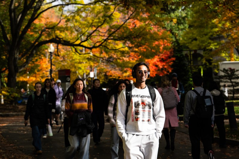 A smiling man walks in a crowd of people with brightly colored foliage behind him.