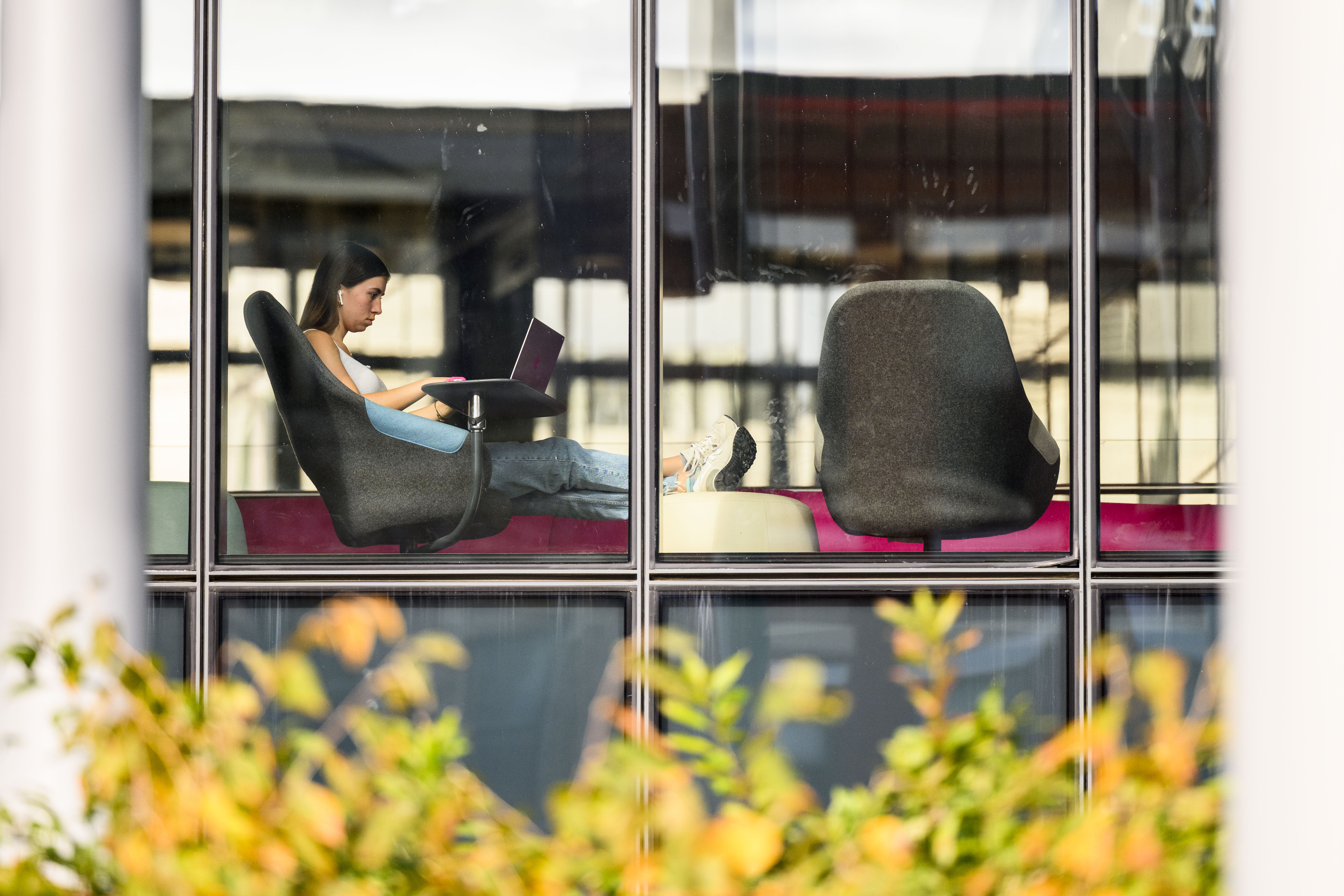 Through a glass window a woman in jeans and a white tank-top sits with her legs outstretched on a white stool as she works on a laptop.