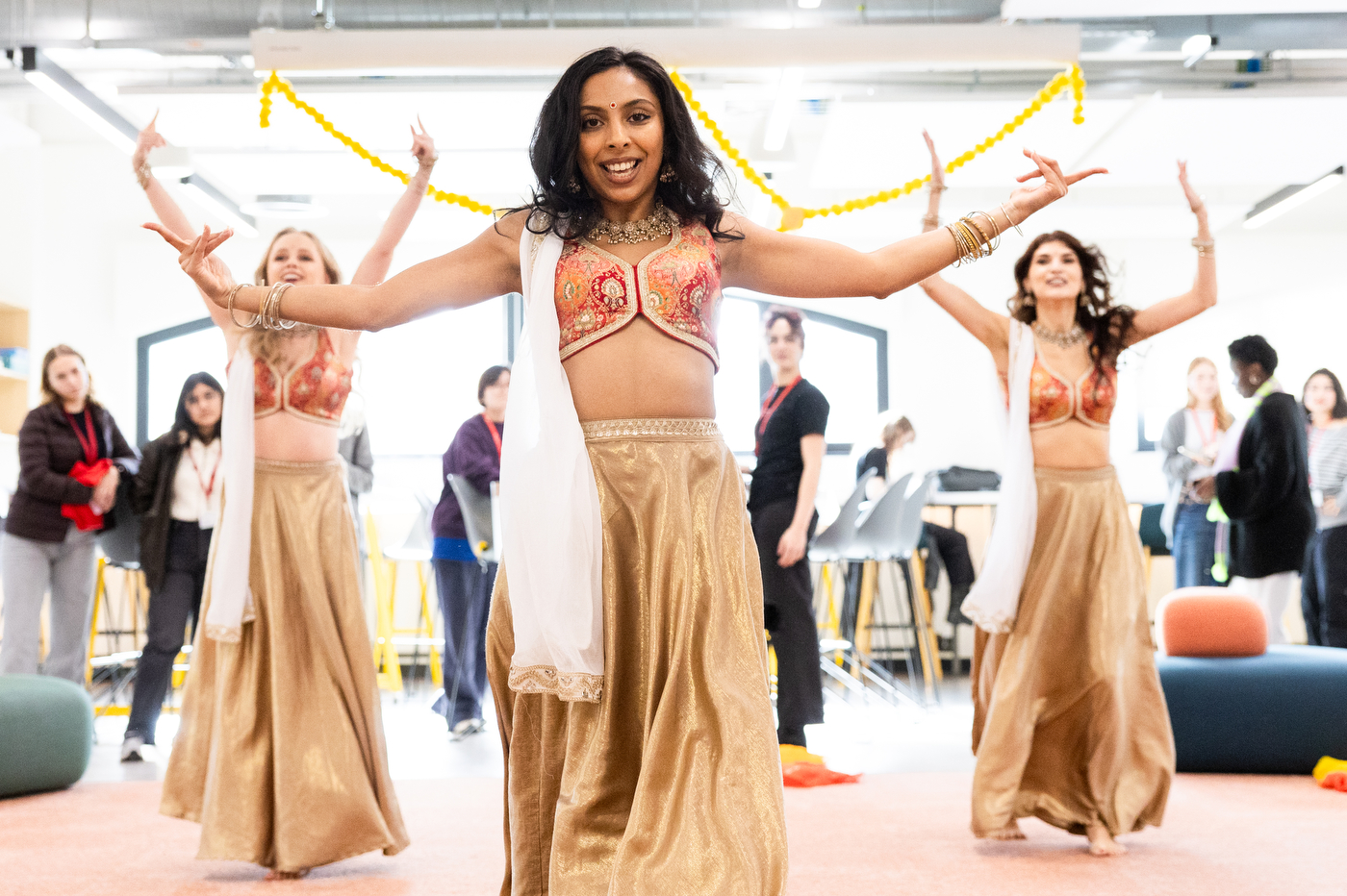 Three dancers in traditional Indian attire perform in a community space at Northeastern University's Devon House. The lead dancer in the foreground wears an ornate coral and gold crop top with a gold skirt and statement jewelry, smiling with arms outstretched. Two dancers behind her mirror the choreography, also wearing matching coral and gold costumes.