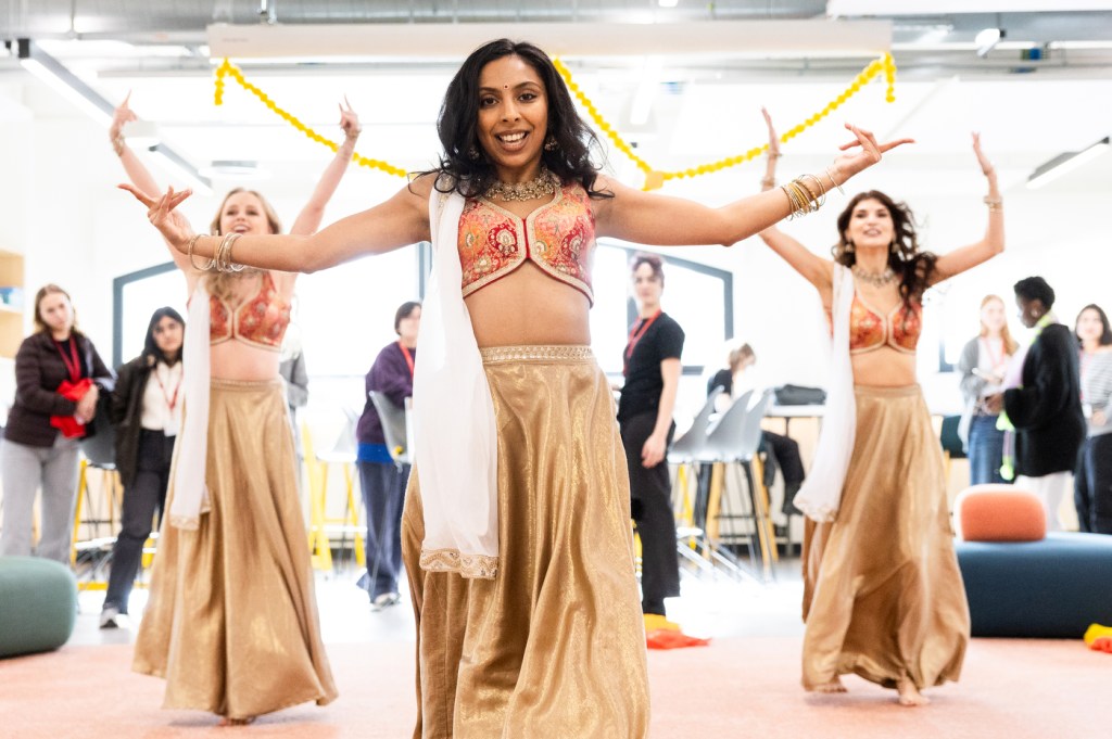 Three Bollywood dancers in gold traditional attire perform with yellow garlands during a Diwali celebration on campus, arms raised in synchronized pose.