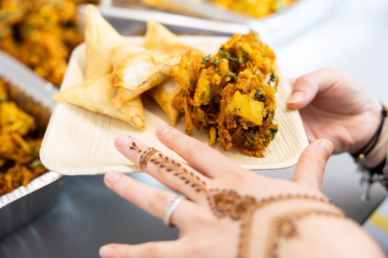 A person with henna designs on their hands holds a small rectangular bamboo plate containing samosas and what appears to be a yellow-orange vegetable dish, possibly pakoras or bhaji. The plate is held over a table where more similar food items are visible in the background. The person is wearing rings and a bracelet, and the henna pattern is visible on their palm and fingers.