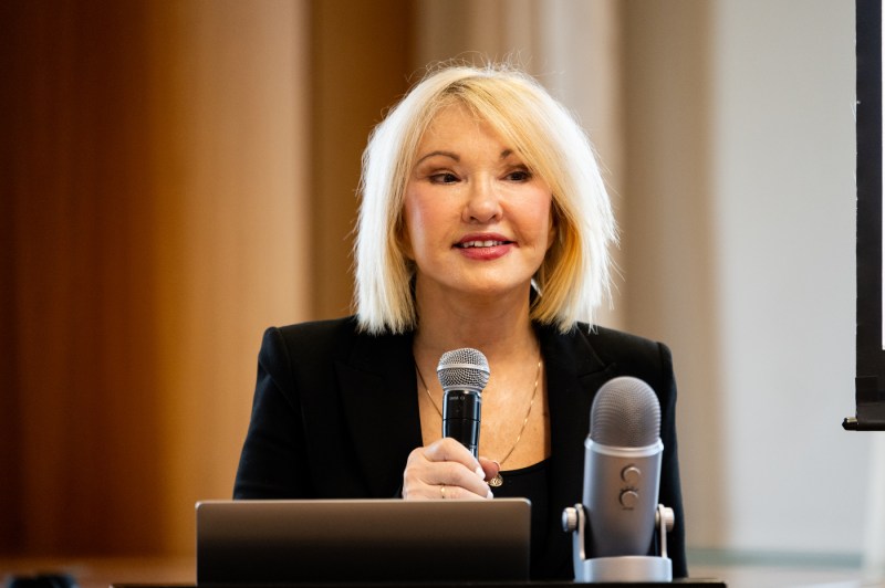 A woman with a blonde bob in a black suit holds a microphone as she speaks at a podium.