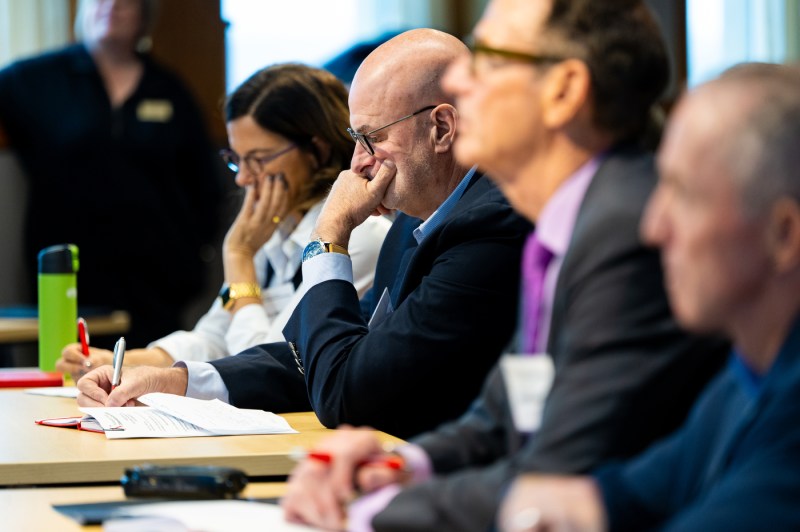 A group of business professionals seated in a row judge a speaker. The focus is on a bald man wearing glasses and a dark suit who is thoughtfully resting his chin on his hand while taking notes. He is writing in a notebook with a pen. Next to him sits a woman in professional attire, and several other attendees are visible but out of focus in the background.